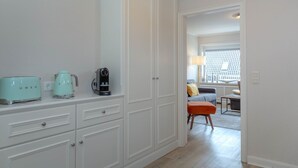 A hallway with a kitchenette area featuring a toaster, kettle, and coffee maker. There is a door leading to a living area with a sofa and a lamp. - Strand Loft - Ferienwohnung "strand-loft" in Westerland (Sylt)