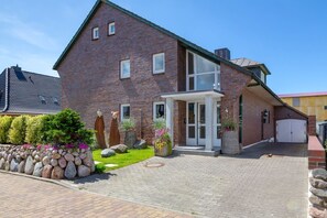 Une maison à deux étages, construite en briques, avec un porche à l’avant, un garage et un jardin bien entretenu agrémenté de rochers et de plantes.