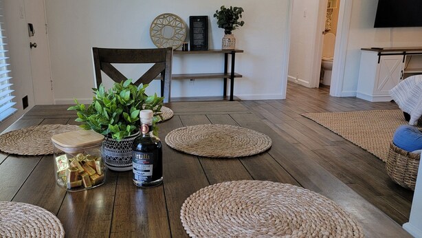 A dining area with a wooden table, woven placemats, a potted plant, a bottle of wine, and a decorative jar.