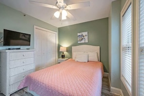 A bedroom with a bed, a ceiling fan, a television, a dresser, and a window with blinds.