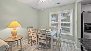 A dining area with a table and chairs, a ceiling fan, a window with blinds, and a refrigerator.