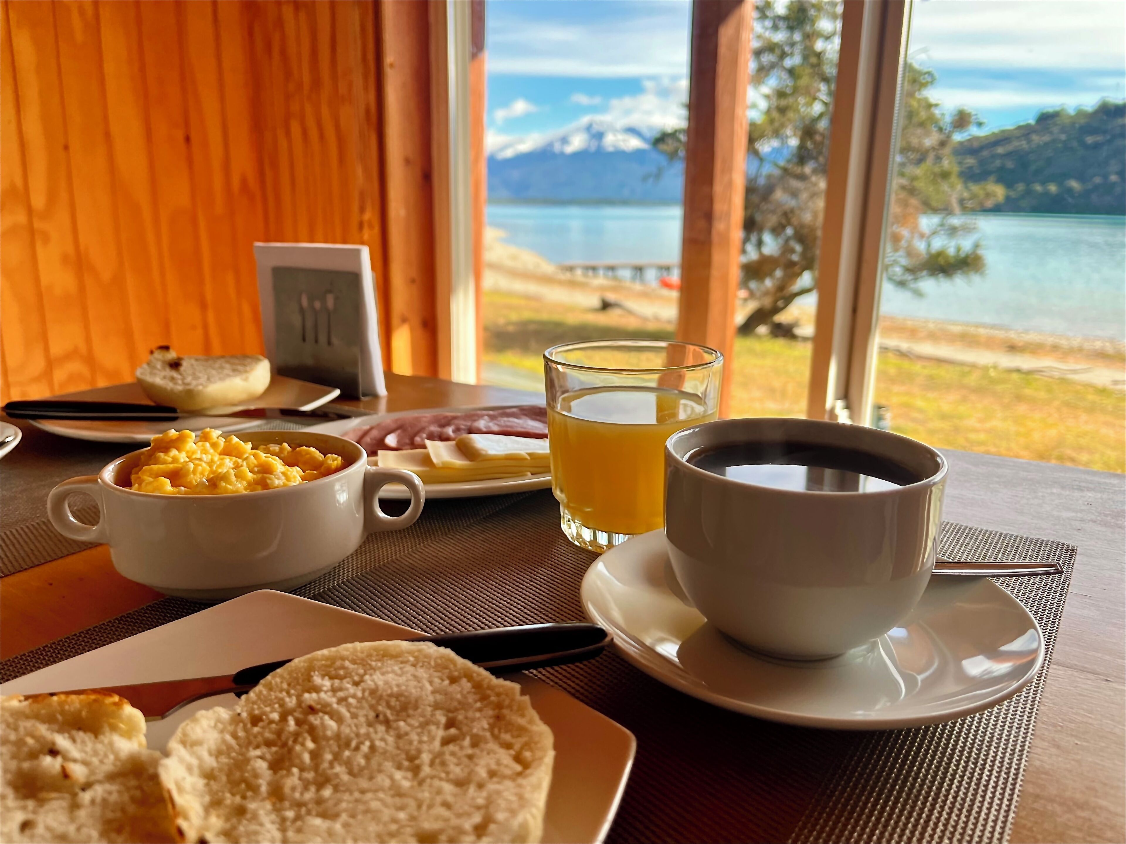 Una mesa de desayuno con huevos revueltos, tostadas, jamón y jugo, con vista a un lago y montañas.