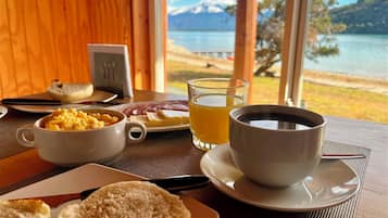 A breakfast table with scrambled eggs, toast, ham, and juice, overlooking a lake and mountains.