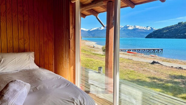A bedroom with a large bed, wooden walls, and a view of a lake and mountains. - Patagonia 47g (Chile Chico)