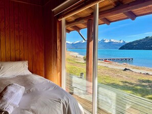 A bedroom with a large bed, wooden walls, and a view of a lake and mountains. - Patagonia 47g (Chile Chico)