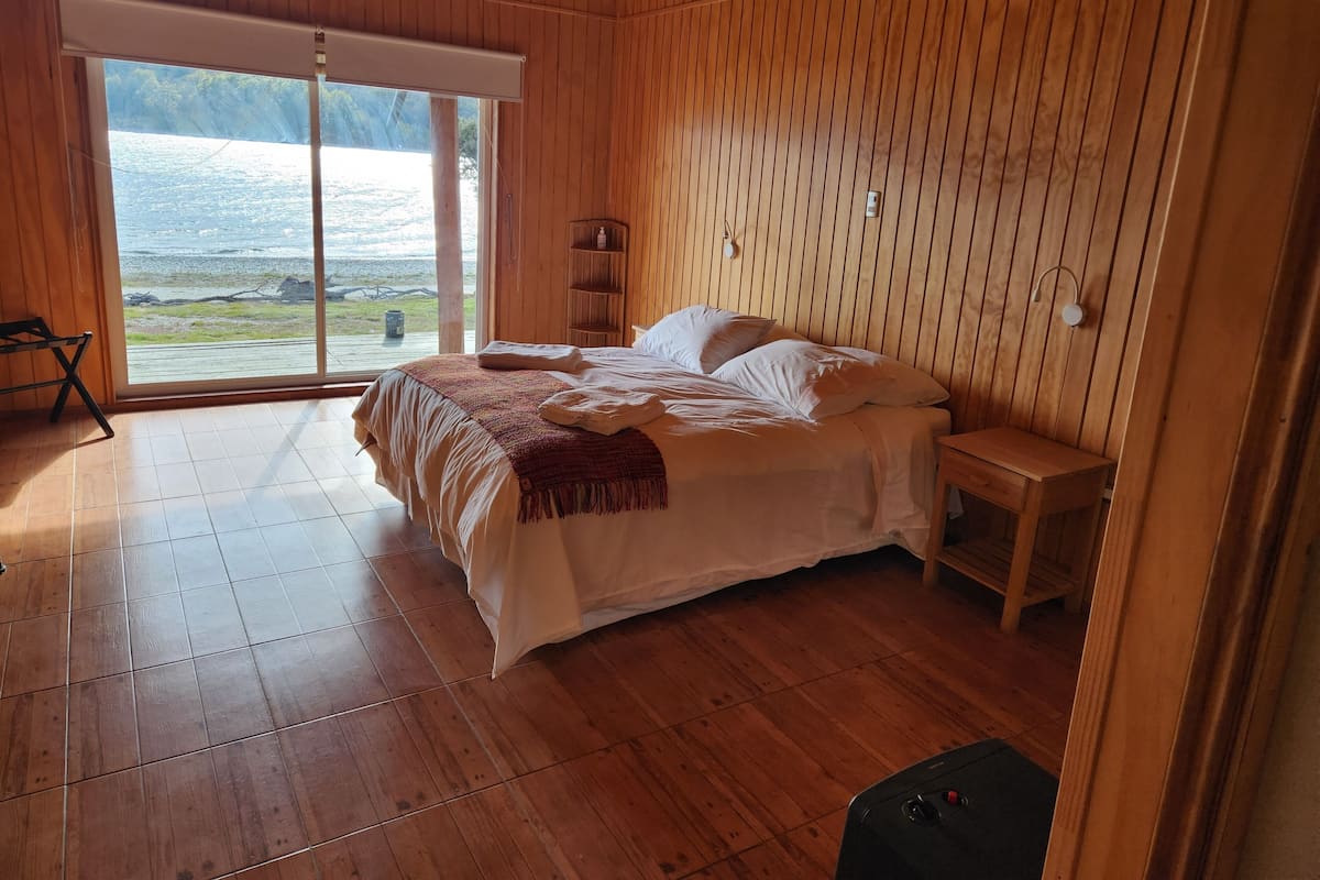 A wooden-paneled bedroom with a bed, a bedside table, and a view of the beach through a large window.