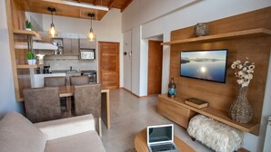 A modern living room with a sofa, a coffee table with a laptop, a TV mounted on a wooden wall, and a kitchen area in the background. - BELGRANO SUITE (San Carlos de Bariloche)