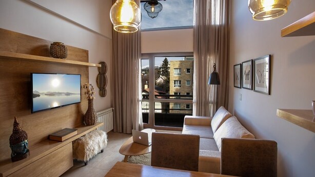 A modern living room with a flat-screen TV, wooden furniture, a laptop, and a view of a building through large windows. - BELGRANO SUITE (San Carlos de Bariloche)