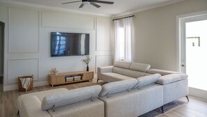Ein modernes Wohnzimmer mit einem großen Ecksofa, einem flachbildfernseher an der Wand, einem Holzkonsolentisch und einem großen Fenster mit Vorhängen.