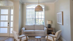 Ein Wohnzimmer mit einem beigefarbenen Sofa, zwei weißen Sesseln, einem runden Couchtisch, einer hängenden Lampenschirm aus Gewebe, einem Beistelltisch mit einer Lampe und einem gerahmten Bild an der Wand.