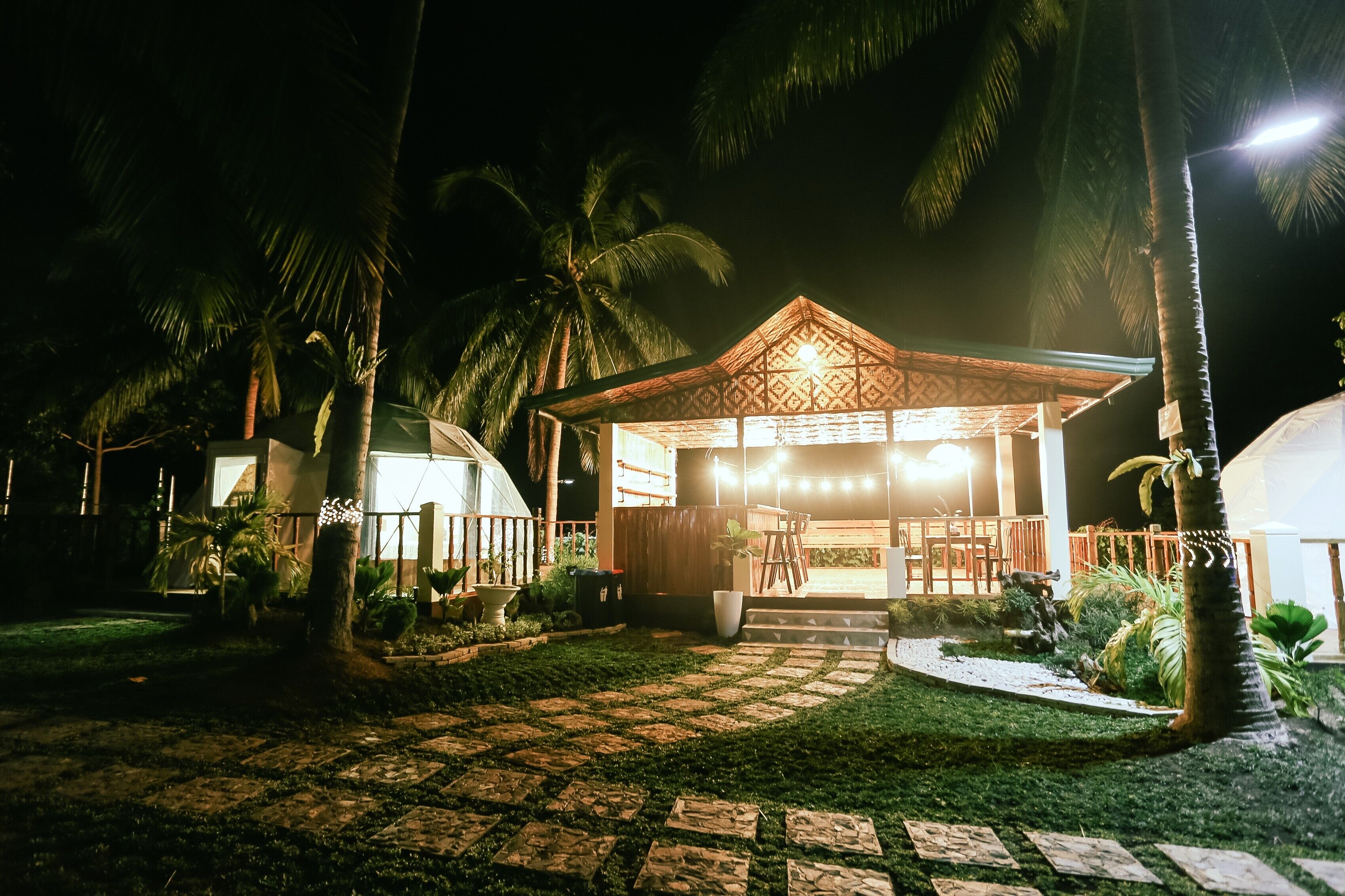 A well-lit gazebo with a patterned roof, surrounded by palm trees and a paved pathway.