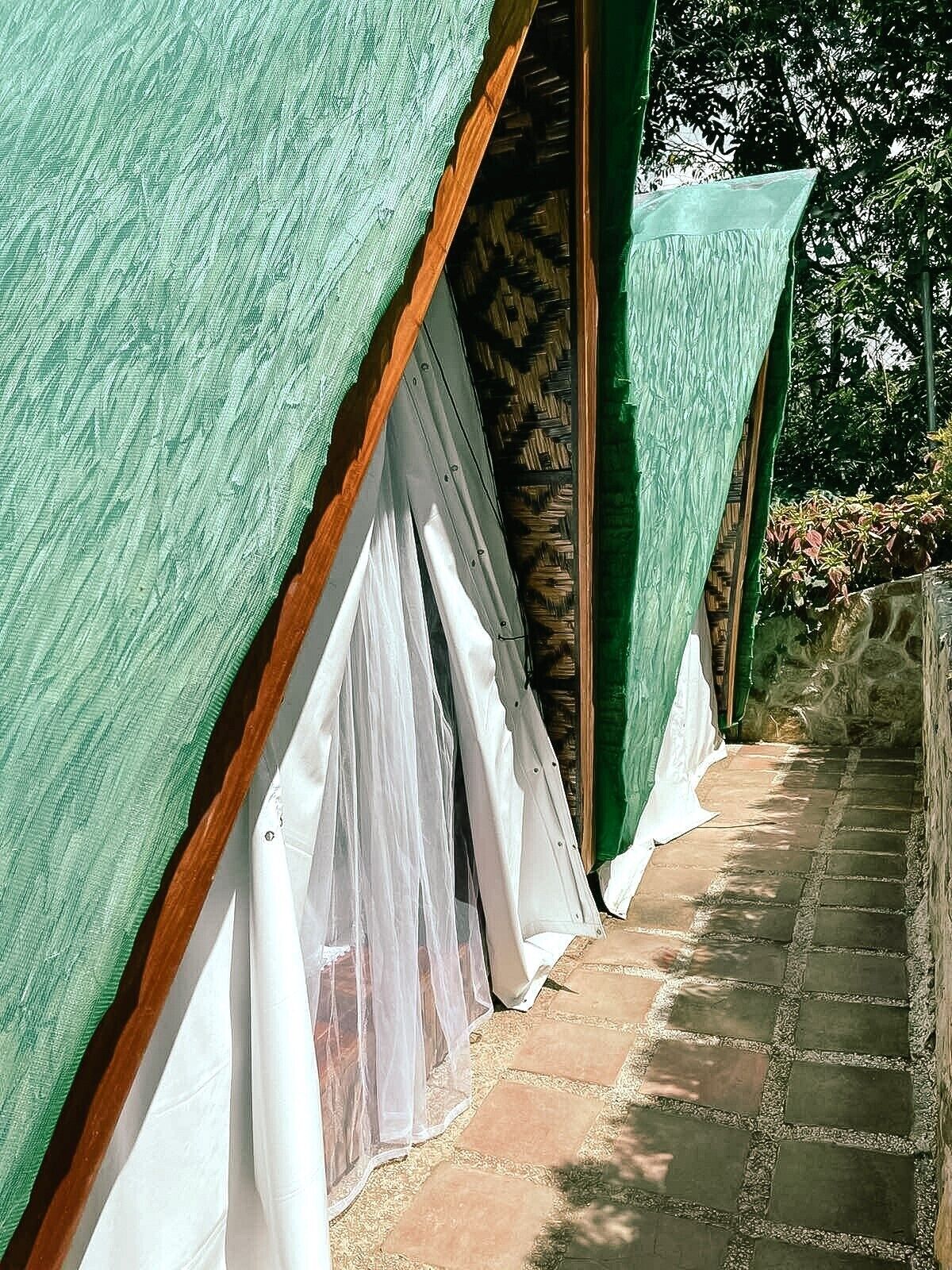 A row of tents with green tarps and white curtains, set against a stone pathway and surrounded by greenery.