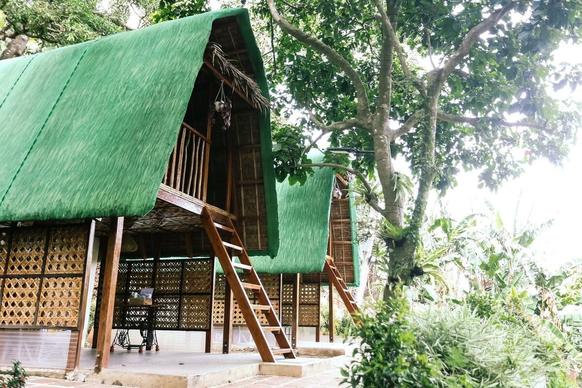 A traditional wooden structure with a green thatched roof, wooden stairs, and a lattice wall.