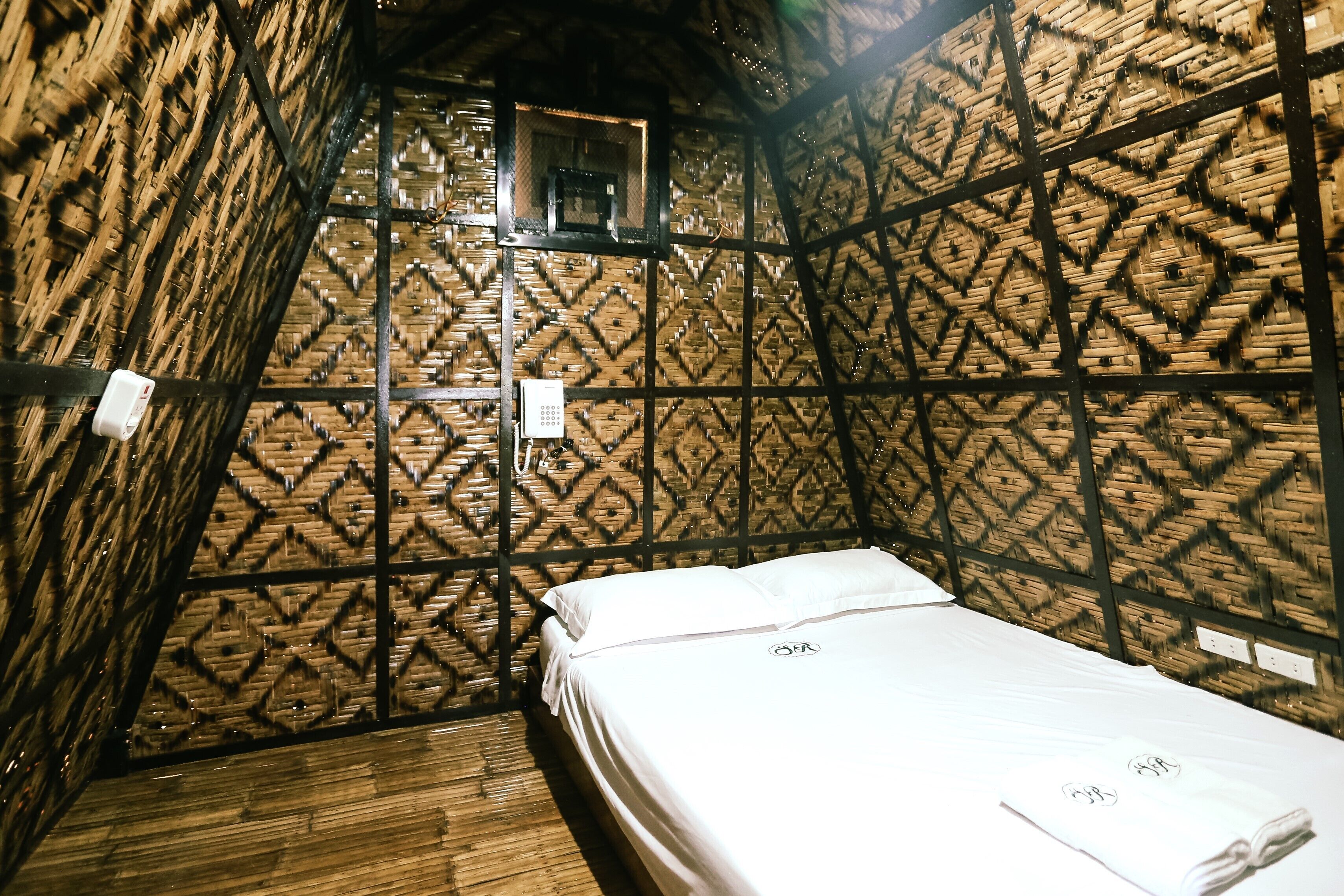 Ein kleiner, gemütlicher Raum mit einem Bett, einer Holzwand mit geometrischem Muster und einem kleinen Fenster.