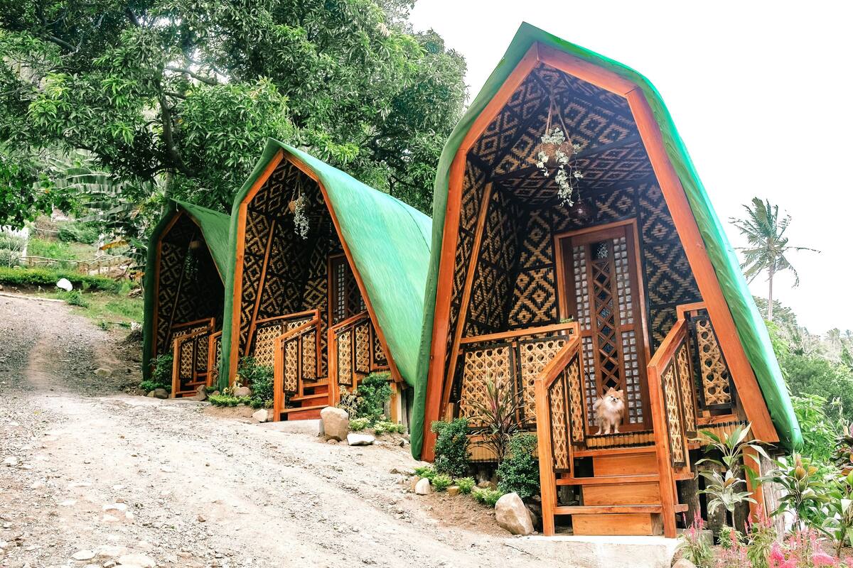 A row of unique, tent-like structures with intricate wooden lattice designs, set on a dirt path surrounded by lush greenery.