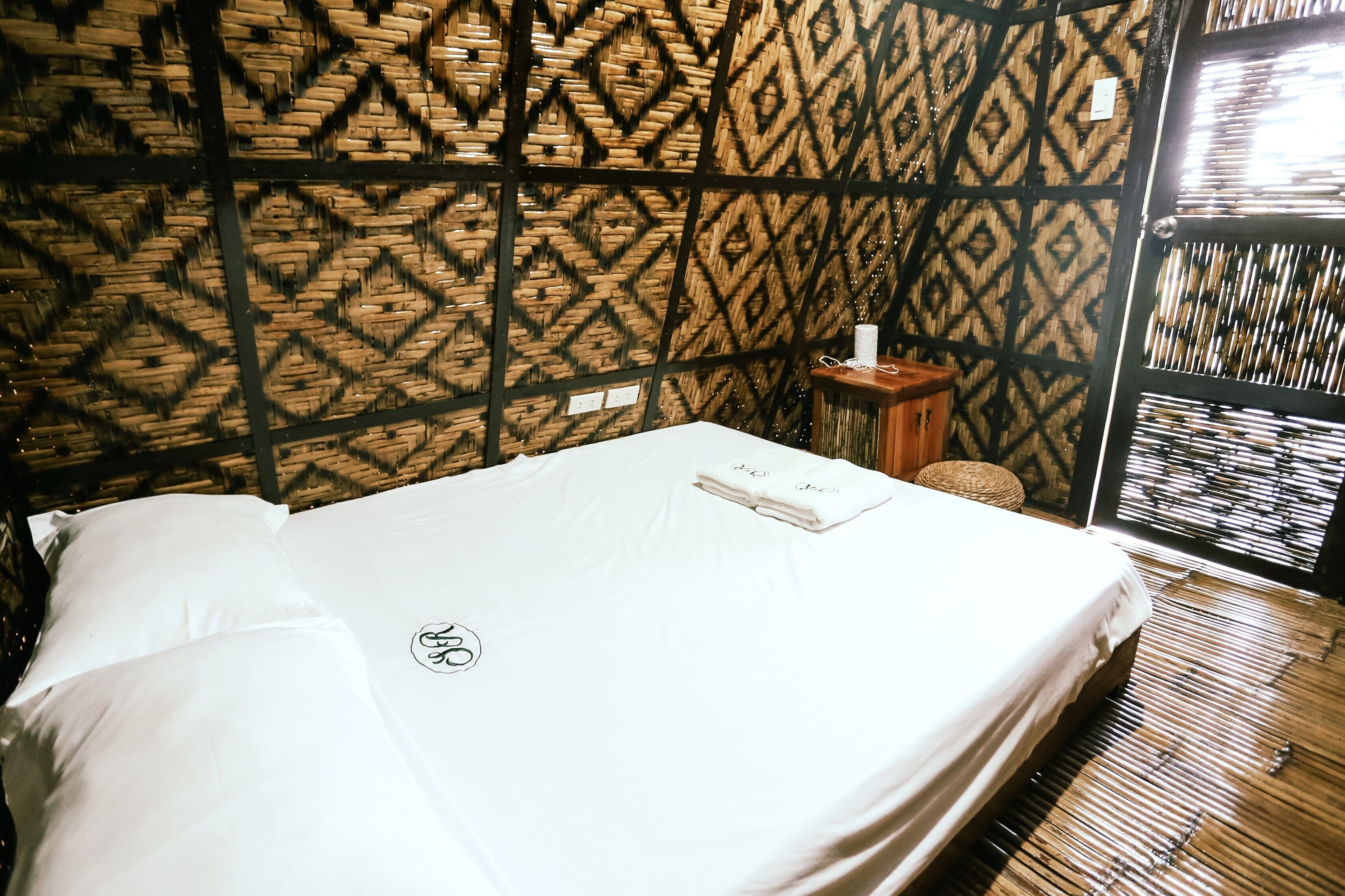 A neatly made bed with a white sheet and a towel, a small wooden side table with a tissue box, and a patterned wall with a geometric design.