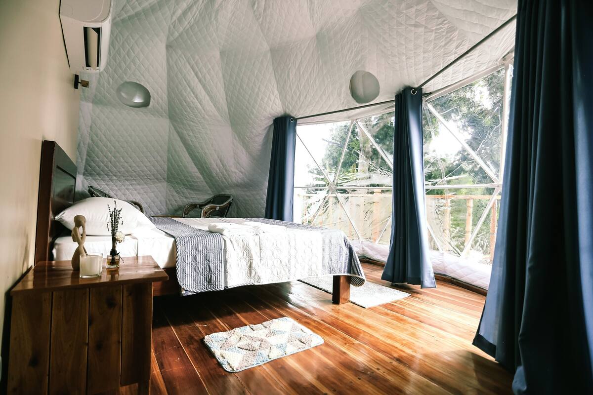 A bedroom with a bed, wooden nightstand, and a view of the outdoors.