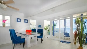 Un comedor moderno con mesa de vidrio y sillas azules, ventilador de techo y vista al mar a través de amplias ventanas.