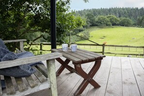 A wooden deck with a picnic table and a bench, overlooking a green landscape with trees and a fence.