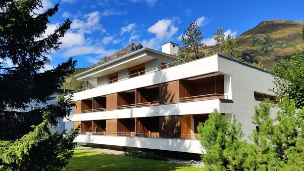 A modern two-story building with balconies, surrounded by greenery and mountains.