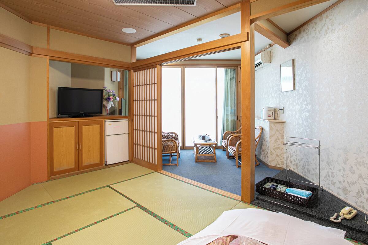 Une pièce de style japonais traditionnel, avec un sol recouvert de tatamis, une télévision, un réfrigérateur et une porte coulissante donnant sur un balcon.
