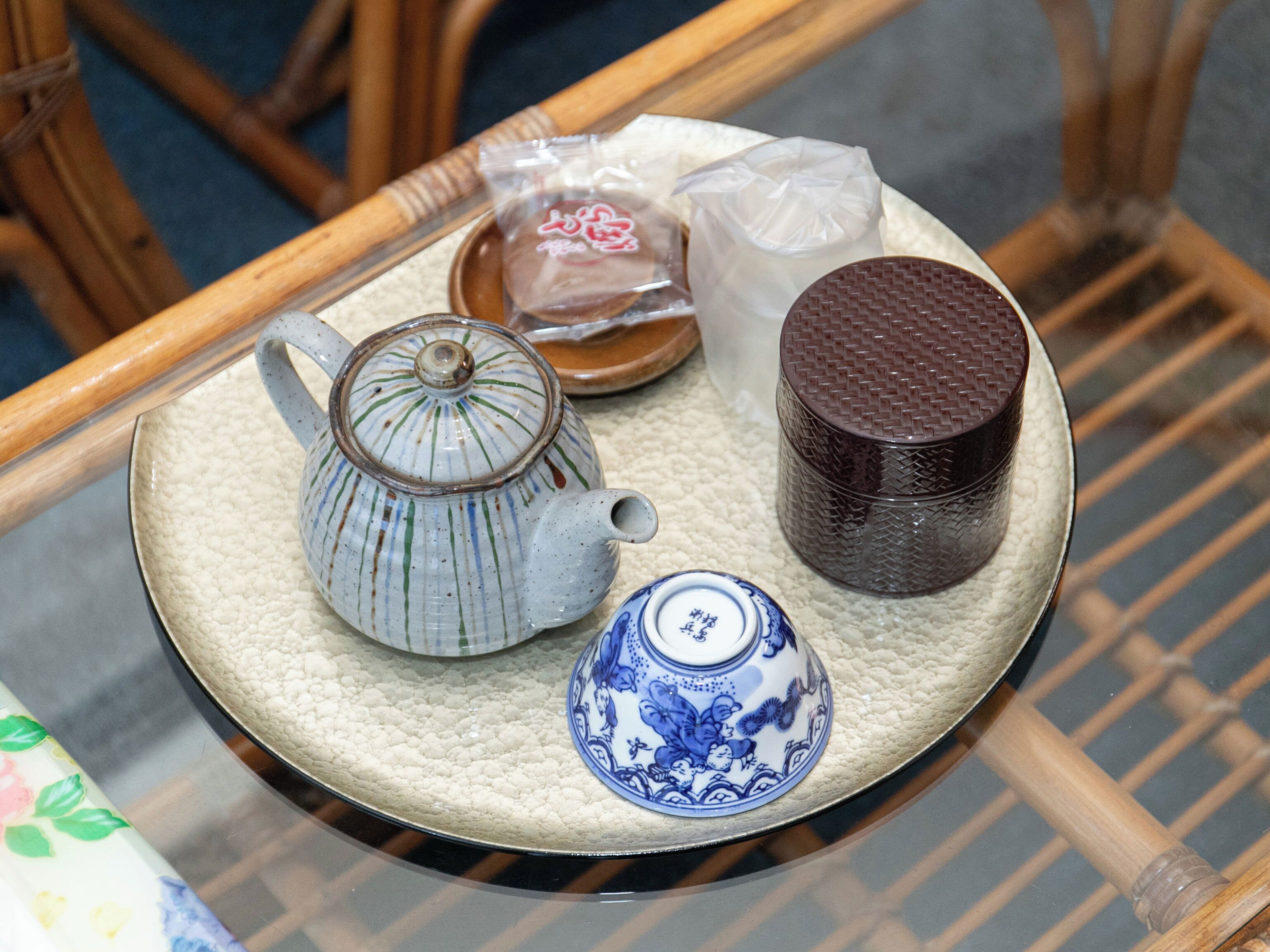 A tray with a ceramic teapot, a blue and white cup, a brown container, and a plastic bag on a woven mat.