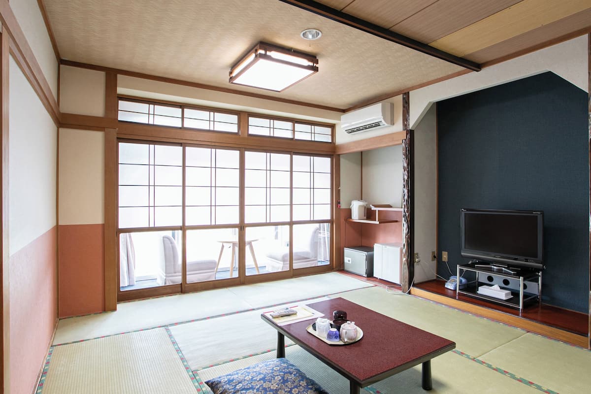 Une pièce de style japonais traditionnel, avec un sol recouvert de tatamis, une petite table avec un coussin, un téléviseur et une porte coulissante donnant sur un balcon.