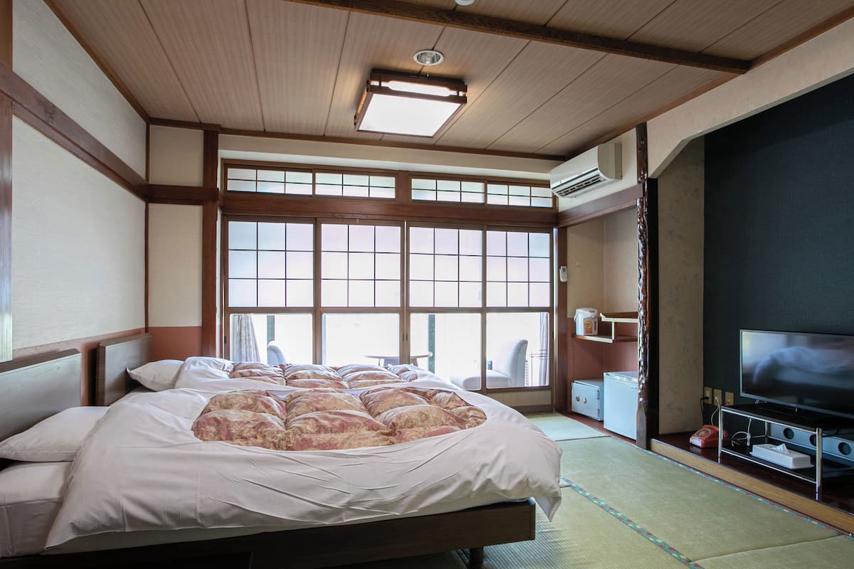 Une chambre de style japonais traditionnel, avec un sol recouvert de tatamis, un grand lit avec une couverture à motifs floraux, une télévision et une porte coulissante donnant sur un balcon.