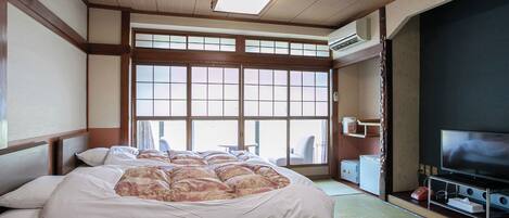 Ein traditioneller japanischer Raum mit Tatami-Boden, einem großen Bett mit floralem Muster, einem Fernseher und einer Schiebetür, die auf einen Balkon führt.