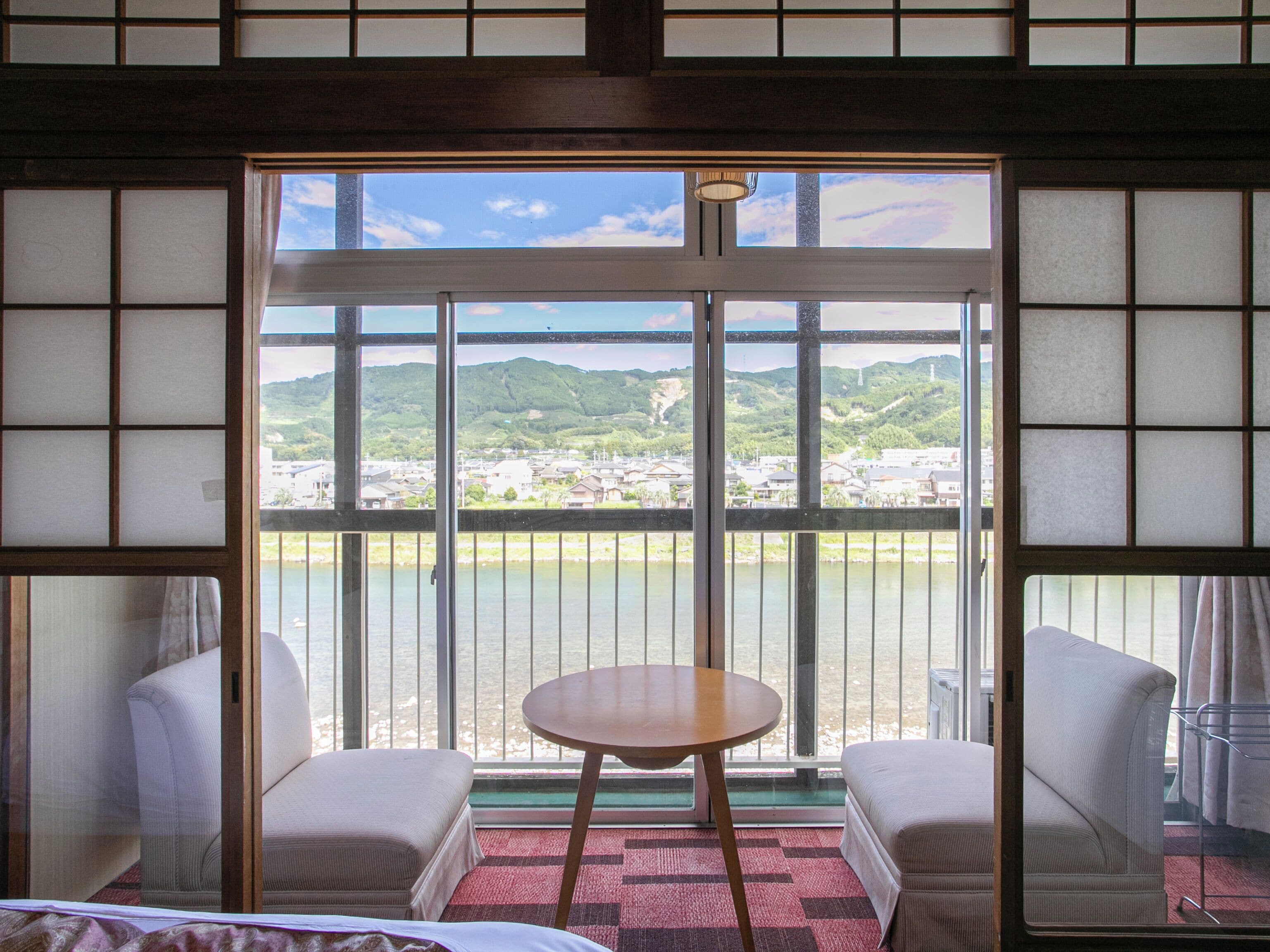 Habitación de hotel con un ventanal que ofrece vistas a un río y colinas distantes. Hay una mesa redonda y dos sillas en la habitación.
