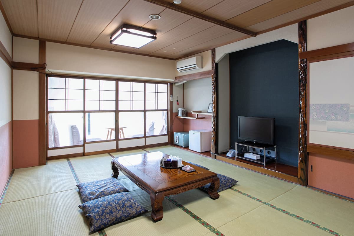 Une pièce de style japonais traditionnel, avec un sol recouvert de tatamis, une petite table en bois, des coussins, un téléviseur et une grande fenêtre.