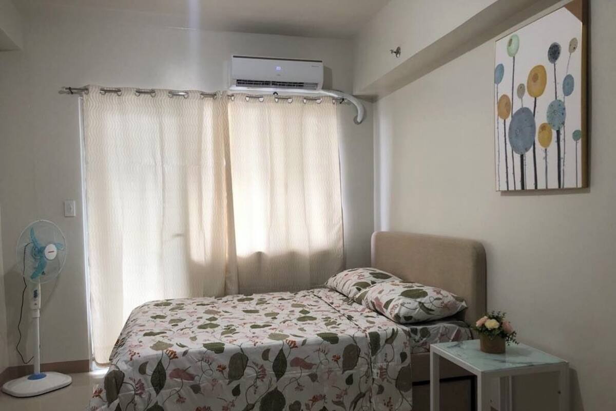 Quarto com cama, mesa de cabeceira com um vaso, ventoinha e janela com cortinas.