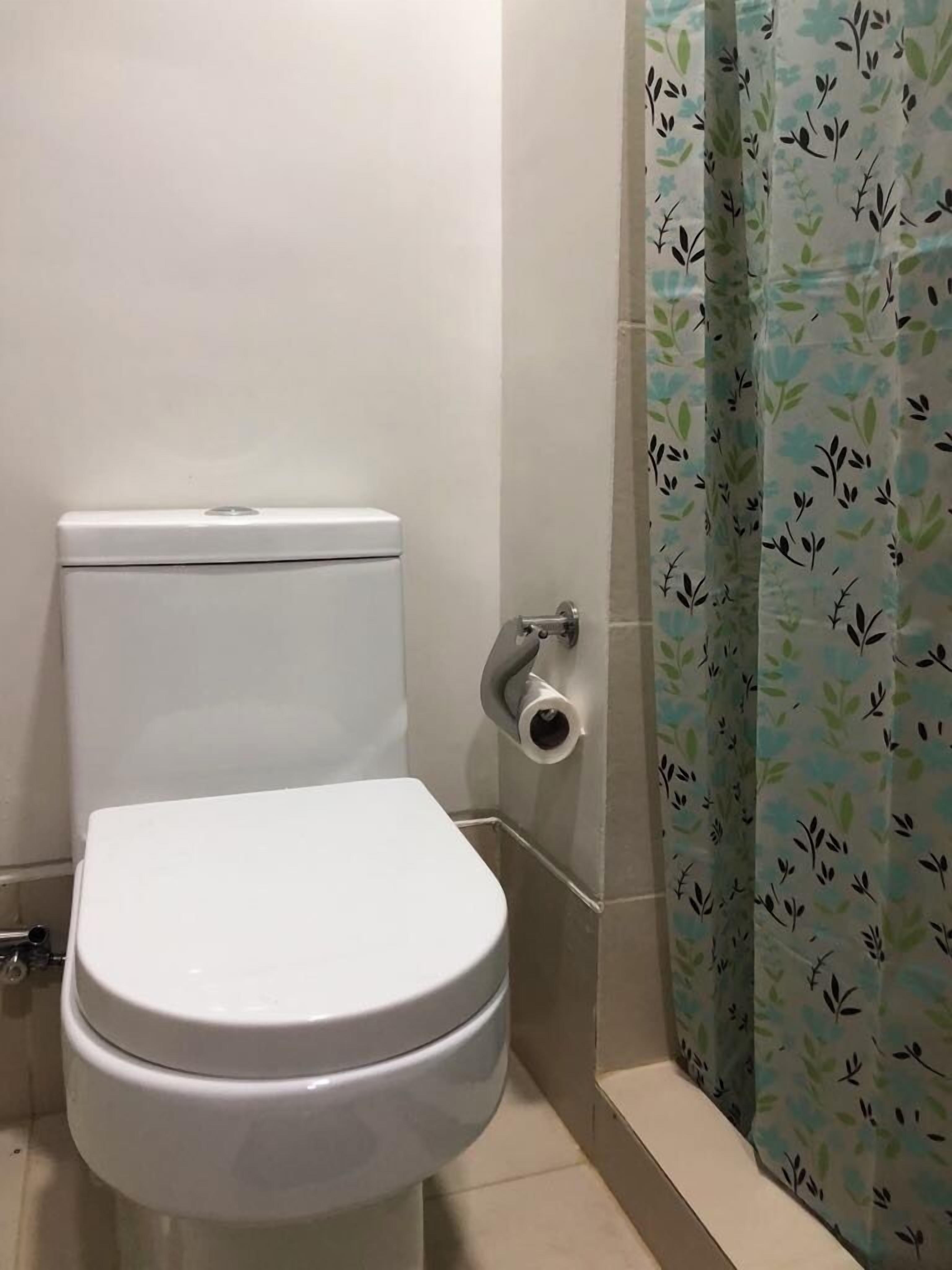 A white toilet with a paper towel holder, a toilet paper roll, and a shower curtain with a leafy pattern.
