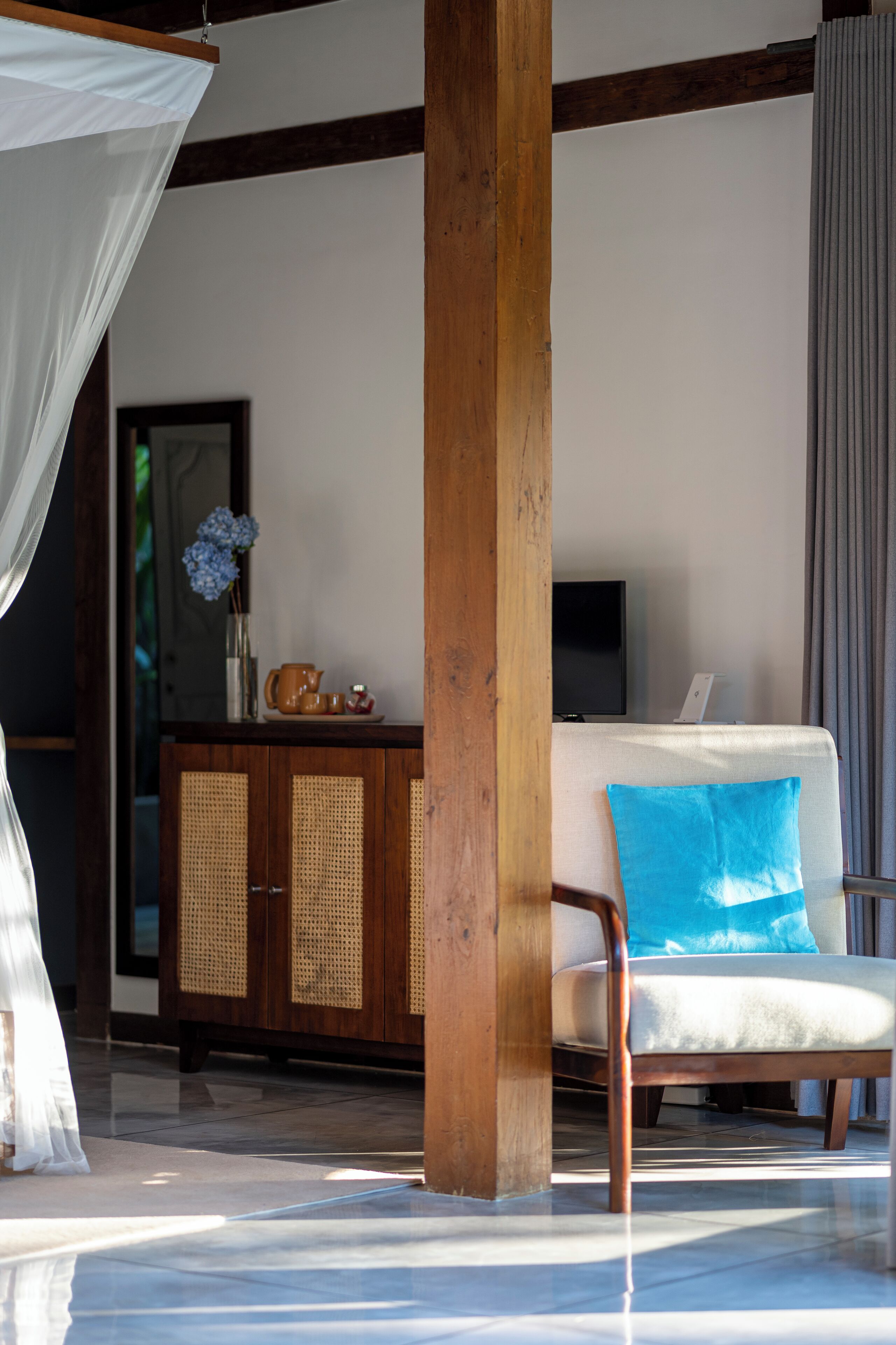 Une pièce avec un pilier en bois, un canapé blanc avec un coussin bleu, un téléviseur posé sur un meuble et un buffet aux portes en osier.