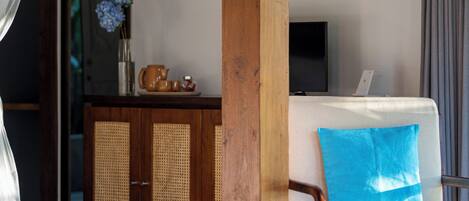 Une pièce avec un pilier en bois, un canapé blanc avec un coussin bleu, un téléviseur posé sur un meuble et un buffet aux portes en osier.