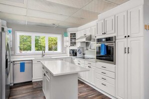 Une cuisine moderne avec des meubles blancs, un îlot central, des appareils en acier inoxydable et une fenêtre donnant sur un espace verdoyant.