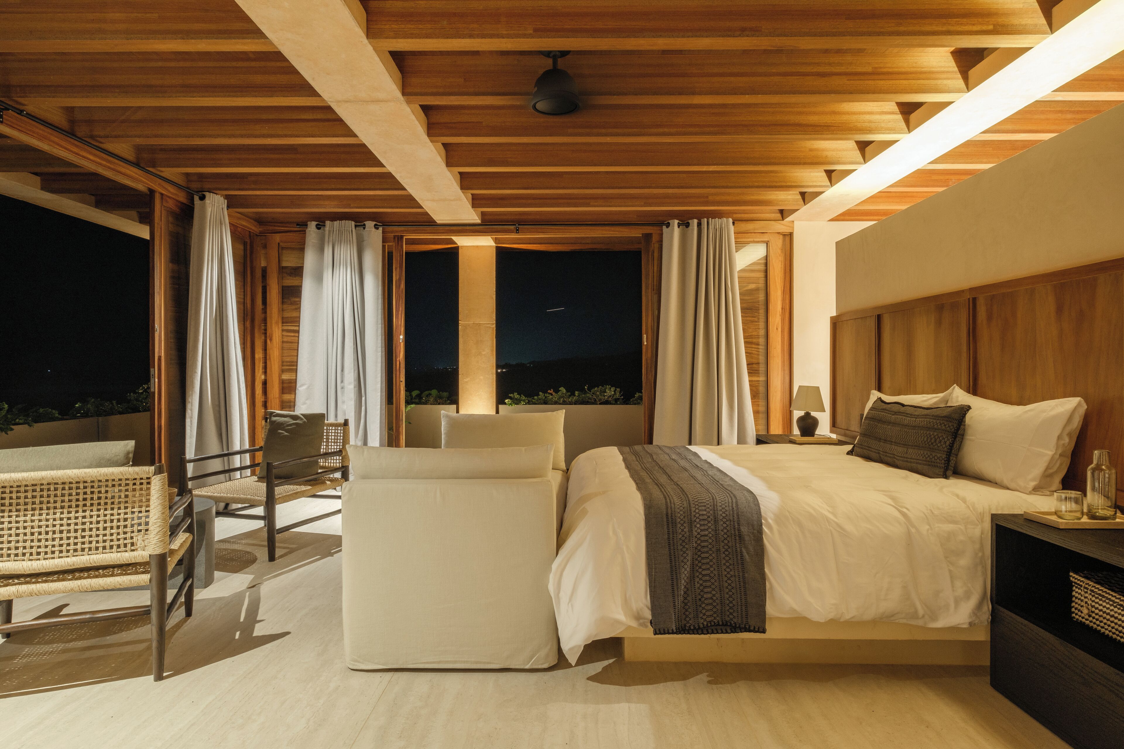 A modern bedroom with a large bed, a balcony with wicker furniture, and a view of the night sky.