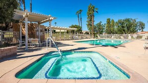 Zona de piscina con área de estar cubierta, palmeras y cielo azul despejado.
