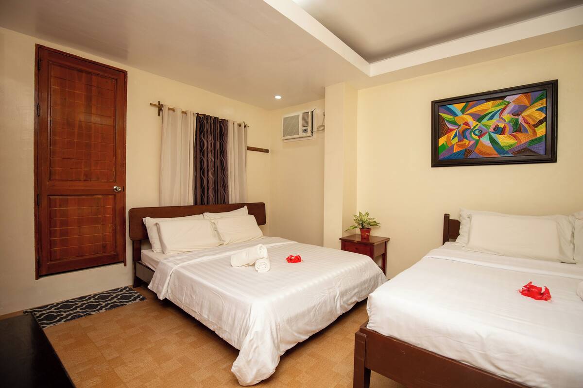 A hotel room with two beds, a wooden door, a window with curtains, an air conditioning unit, and a colorful painting on the wall.