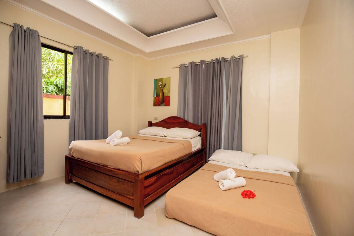 A bedroom with a wooden bed, a smaller bed, grey curtains, a painting on the wall, and a window with curtains.
