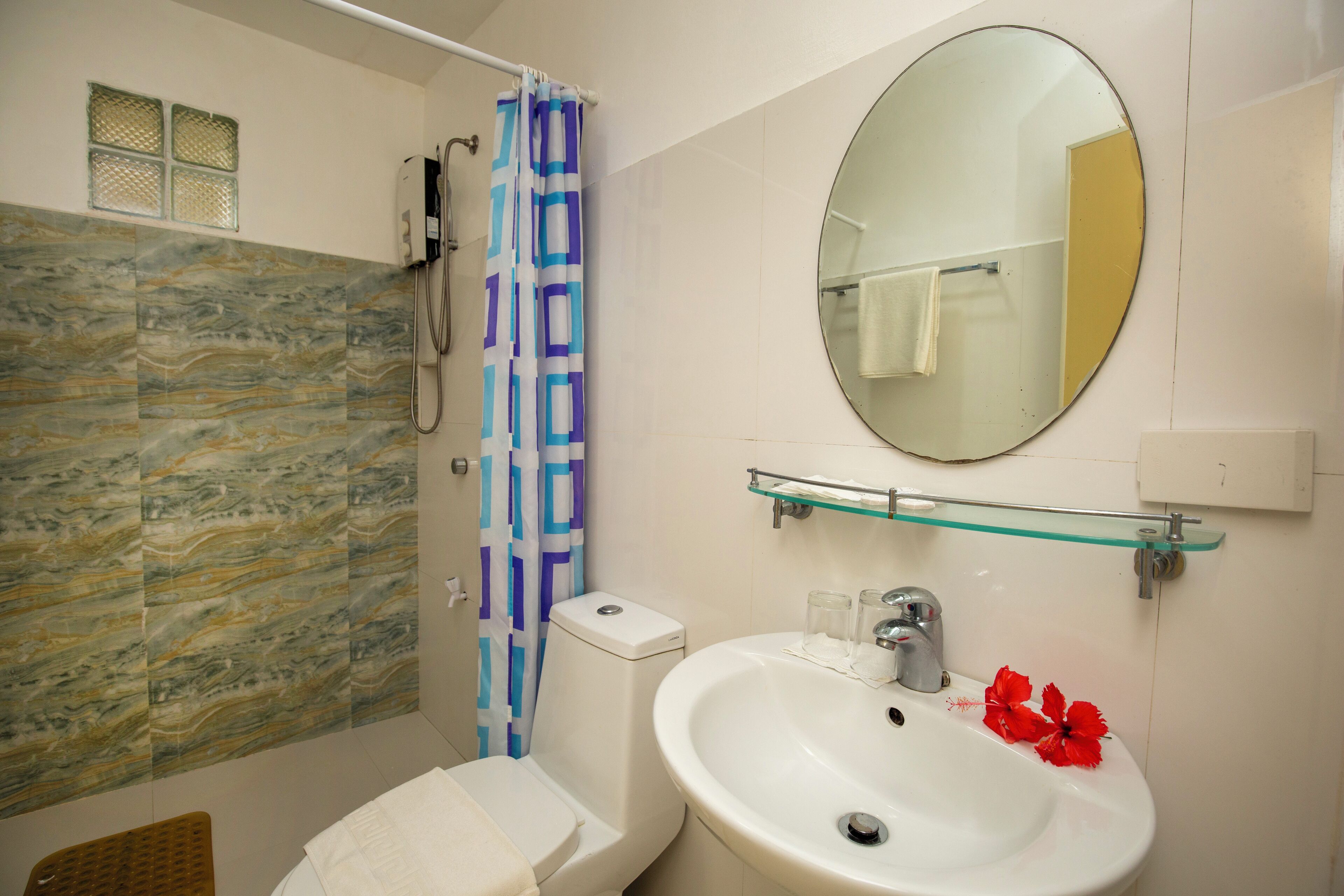 A bathroom with a marble wall, a shower with a blue and white curtain, a white sink with a glass shelf, and a round mirror.