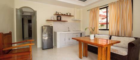A living room with a wooden table and chairs, a sofa, a kitchen area with a refrigerator, and a window with curtains.