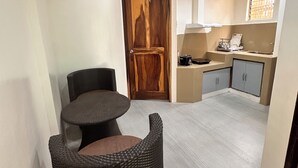 A compact living space with a dining area featuring a round table and a wicker chair, a kitchenette with cabinets, and a wooden door. - Vilus Place  (Boracay Island)