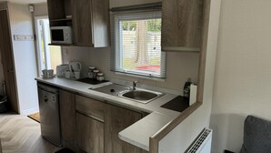 A compact kitchen with wooden cabinets, a microwave, a sink, and a window with blinds. - West Lawn 47 Haggerston Castle Paddock Views (Berwick-upon-Tweed)