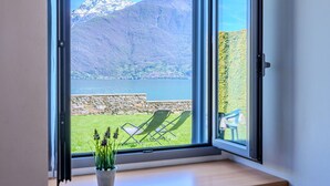 A room with a large window offering a view of a lake, mountains, and a stone wall with outdoor seating. - Beautiful apartment for 4 guests with WIFI, TV and patio (Musso)