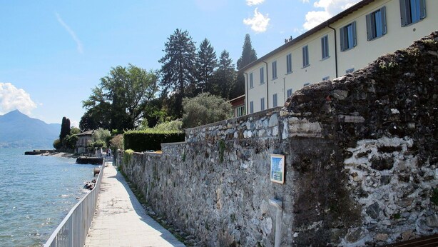 A stone wall with a sign, a paved walkway, and a building with windows overlooking a body of water. - Beautiful apartment for 4 guests with WIFI, TV and patio (Musso)