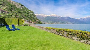 A lakeside view with a grassy area, stone wall, and blue lounge chairs. - Nice apartment for 4 people with WIFI, TV and patio (Musso)