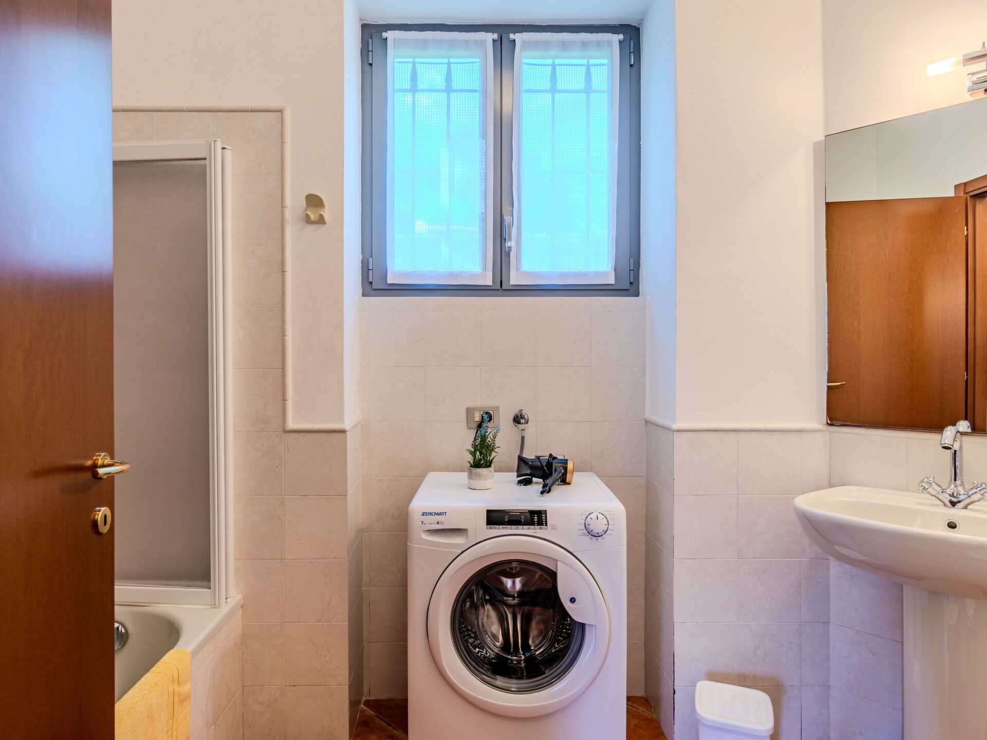 Un baño pequeño con lavadora, lavamanos y ventana sobre el espejo.