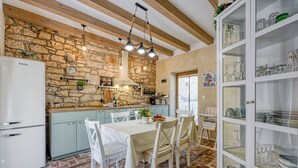 Une cuisine rustique avec un mur en pierre, des poutres en bois et un coin repas comprenant une table et des chaises blanches.