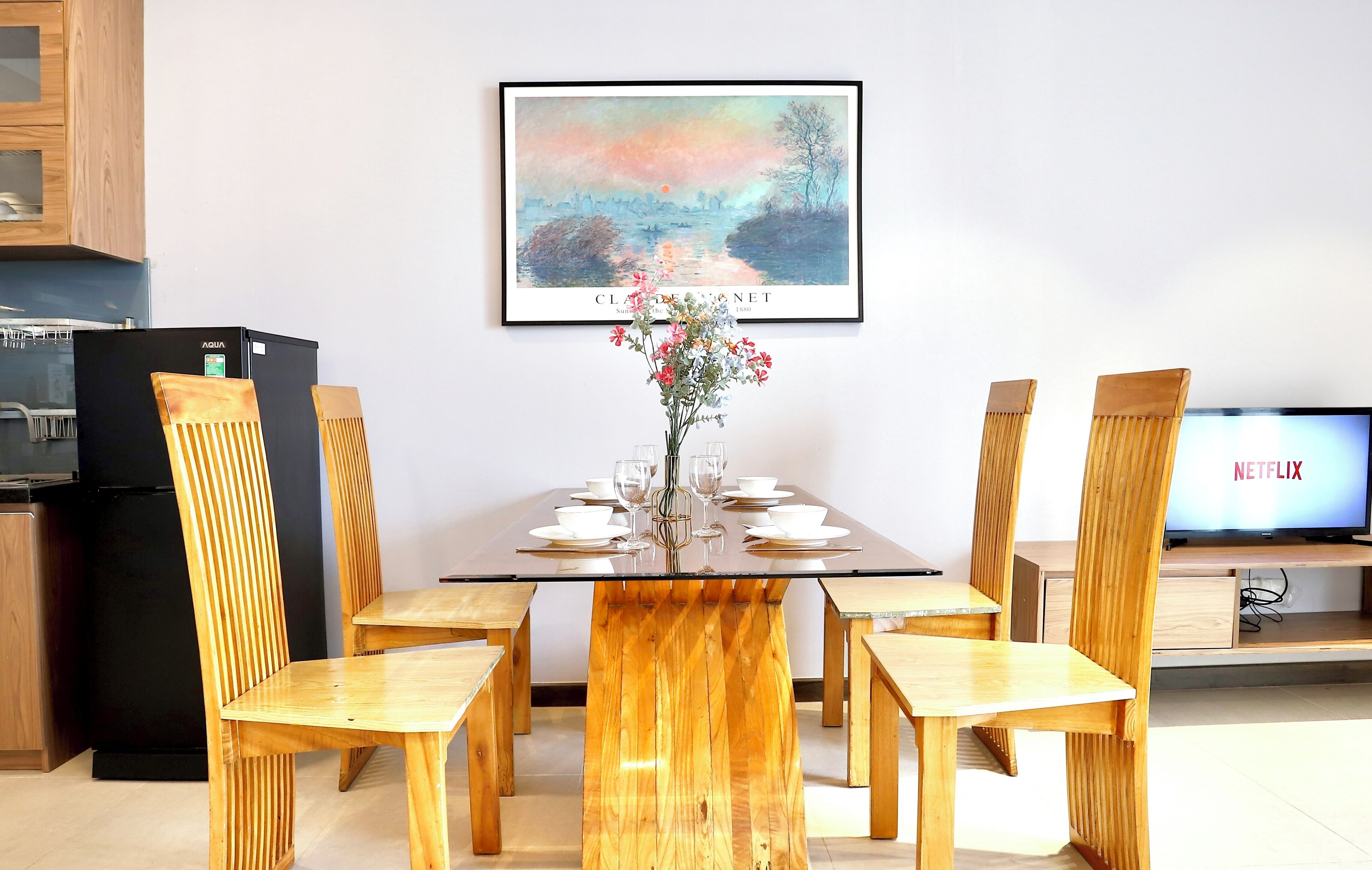 A dining area with a glass-top table, wooden chairs, a vase with flowers, and a framed painting above.